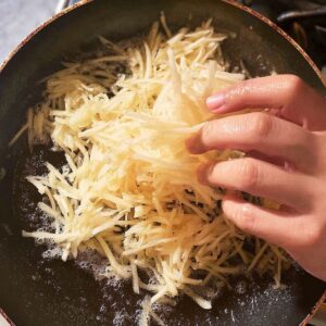 Delicious Crispy Hash Browns Recipe 15 A hand placing shredded raw potatoes into a hot, oiled pan to start cooking hash browns.