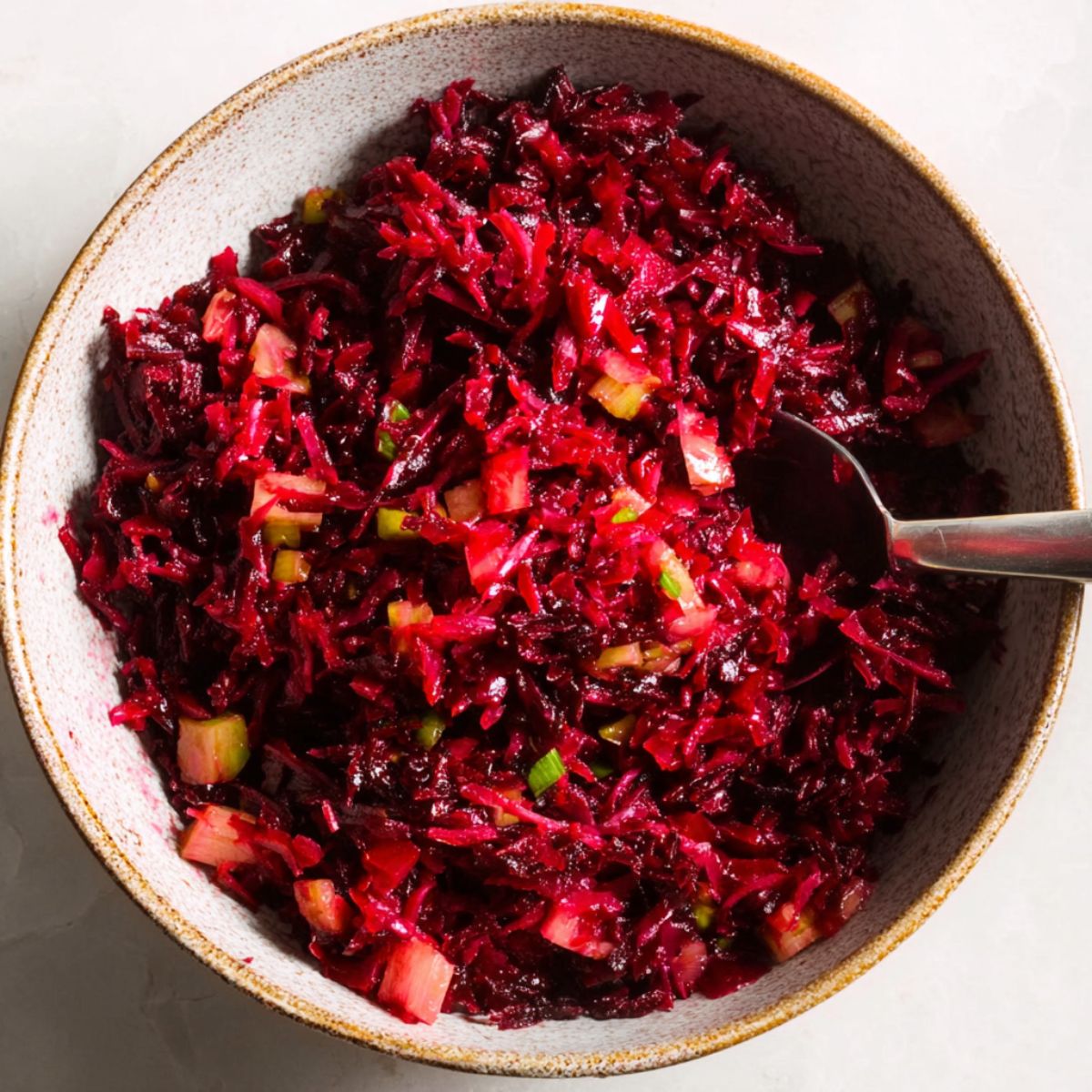 Delicious Grated Beet Salad Recipe 12 A bowl filled with finely shredded or grated beets mixed with small diced vegetables like pickles or apples. The salad has a deep red color from the beets, and a spoon is partially visible in the bowl.