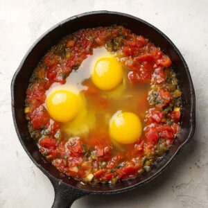 Easy Homemade Shakshuka Recipe 16 Three raw eggs being cracked into the tomato and vegetable mixture in a cast-iron skillet.