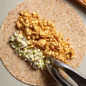Healthy Shredded Tofu Wraps Recipe 18 Overhead view of a whole wheat tortilla being filled with shredded buffalo tofu and a creamy slaw using tongs. The ingredients are placed in a line along the center of the tortilla.