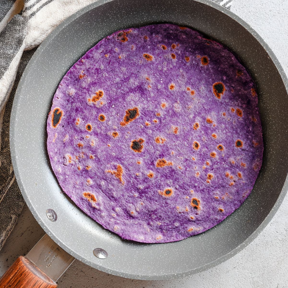 Easy Sweet Potato Tortillas Recipe 12 "Top-down view of a freshly cooked purple tortilla in a gray non-stick skillet, with light char spots indicating it’s perfectly cooked. The skillet has a wooden handle and rests on a neutral textured surface with a striped kitchen towel nearby. Bright natural lighting emphasizes the vibrant purple color and soft texture of the tortilla. Ideal for gluten-free, vegan, or healthy tortilla recipe content.