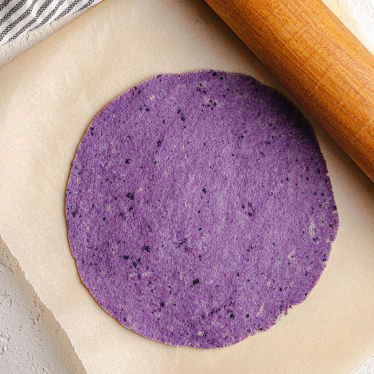 Easy Sweet Potato Tortillas Recipe 11 Top-down view of a freshly rolled raw purple tortilla on parchment paper, with a wooden rolling pin partially visible on the side. The tortilla is vibrant purple with natural speckles and smooth texture. The background features a neutral textured surface with a striped kitchen towel, highlighting the tortilla's color and prepping process. Perfect for gluten-free, vegan, and healthy tortilla recipe content.