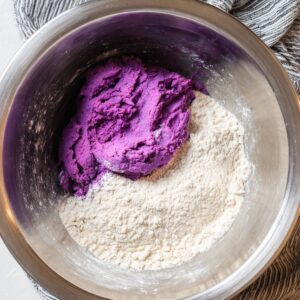 Easy Sweet Potato Tortillas Recipe 14 Top-down view of a stainless steel mixing bowl containing vibrant purple dough and a heap of white flour, ready to be combined for making purple corn tortillas. A striped kitchen towel is draped in the background. The image highlights healthy, gluten-free, and colorful cooking ingredients for vegan and homemade tortilla recipes